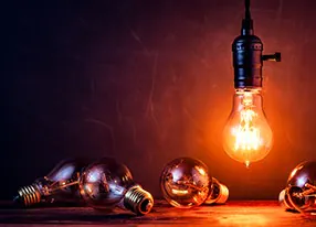 Leuchtende Gl&uuml;hbirne vor einer schwarzen Wand. (iStock.com/ ThomasVogel)